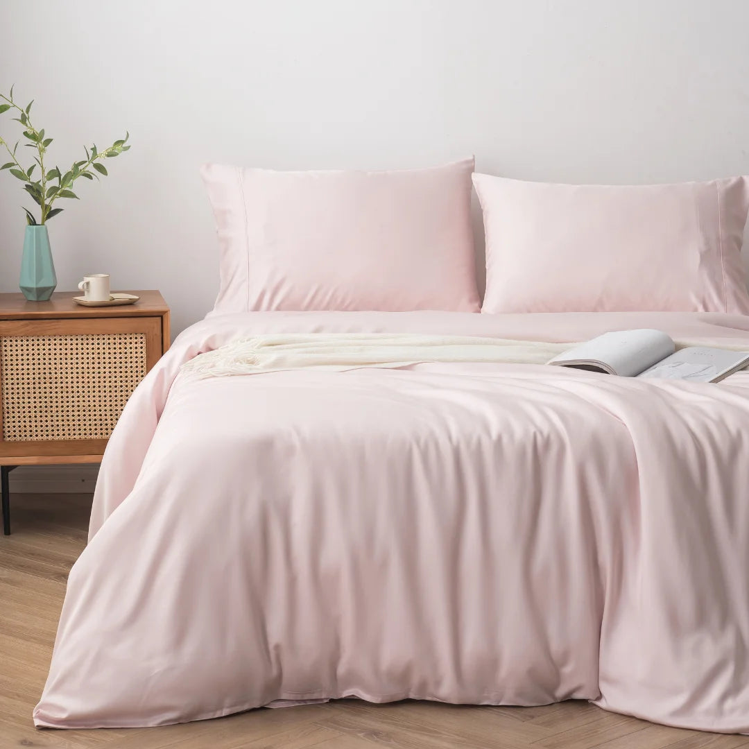 A neatly made bed, adorned with a Linenly Blush Bamboo Quilt Cover for a luxurious sleep experience, and an open book resting on it, beside a nightstand with a vase containing greenery.