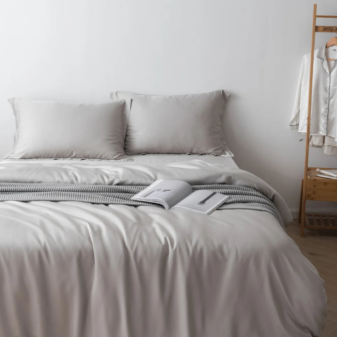 A neatly made bed with luxurious bedding, including a Linenly Silver Bamboo Quilt Cover, a pair of pillows, and an open book lying in the center. A white robe hangs on a wooden rack in the