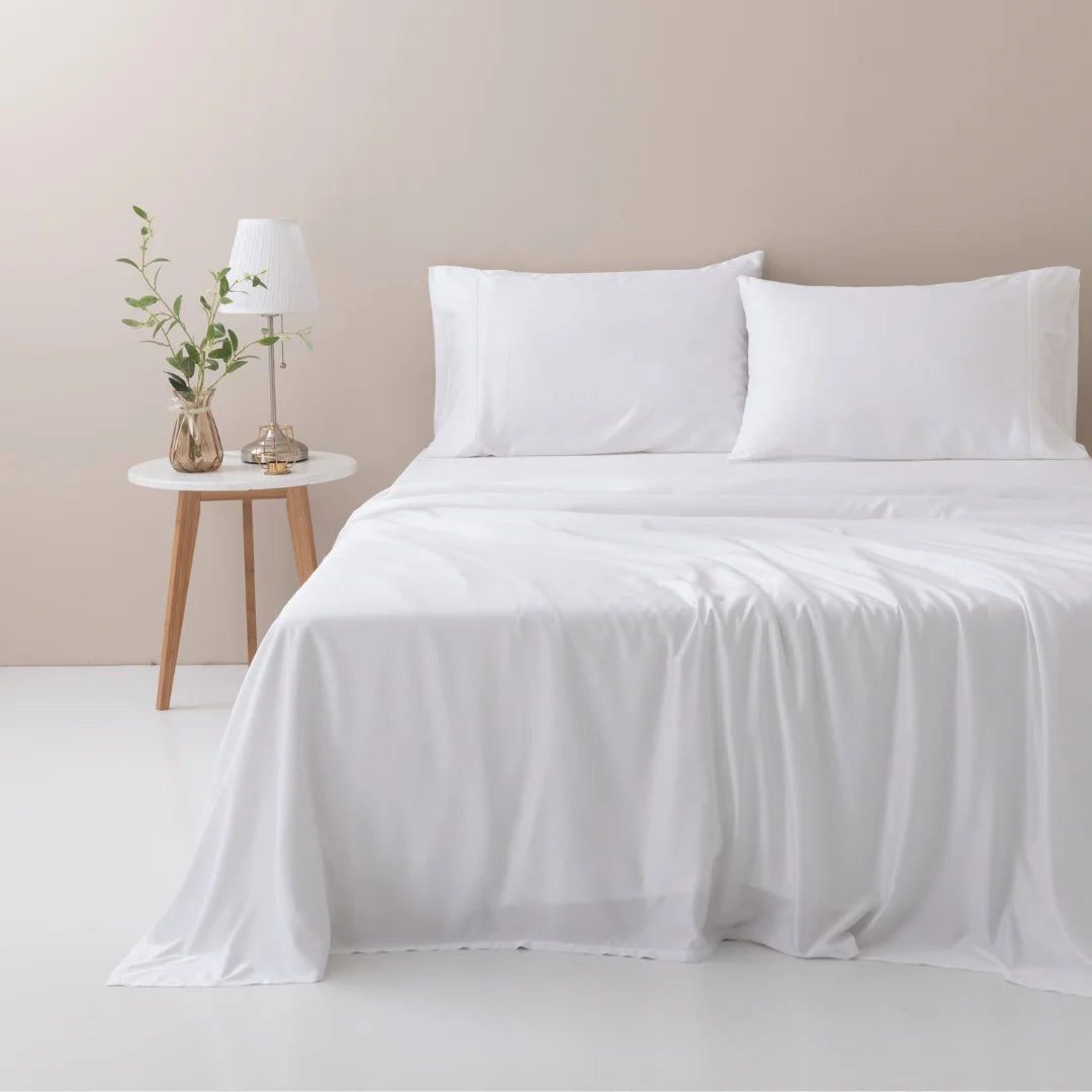 A serene and minimalistic bedroom setup featuring a neatly made bed with Linenly's ultra-soft, white bamboo sheet set, beside a wooden nightstand adorned with a simple lamp and a vase with fresh greenery.