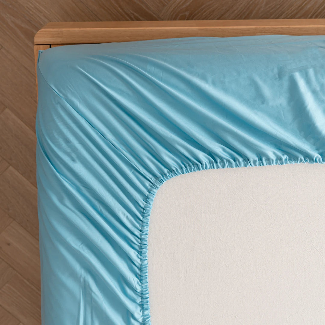 Close-up view of a neatly made bed with a smooth, Linenly aqua blue bamboo fitted sheet draped over the edge of a wooden bed frame, offering a cooling effect.
