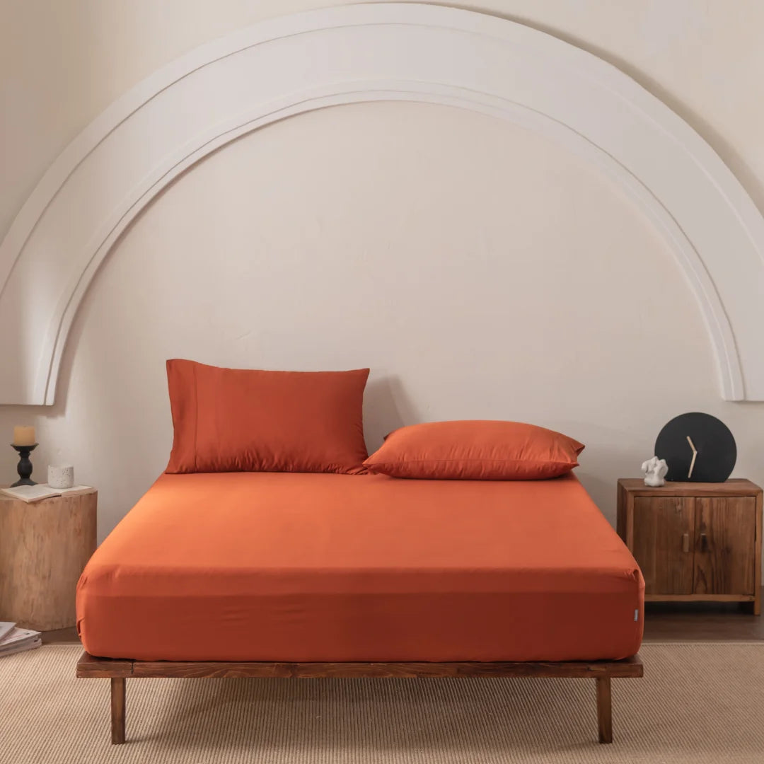 In a minimalist bedroom, comfort is exuded with a bed adorned in orange sheets and pillows. A white arch design serves as a backdrop, while wooden nightstands hold decorative items. Linenly's Bamboo Fitted Sheet in "Copper Sunset" enhances the room's tranquil, neutral palette.