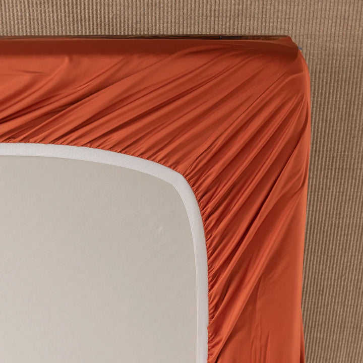 The Bamboo Fitted Sheet - Copper Sunset by Linenly snugly wraps around a beige mattress in a soothing orange hue, creating smooth lines and blending seamlessly with the light brown carpeted floor beneath.