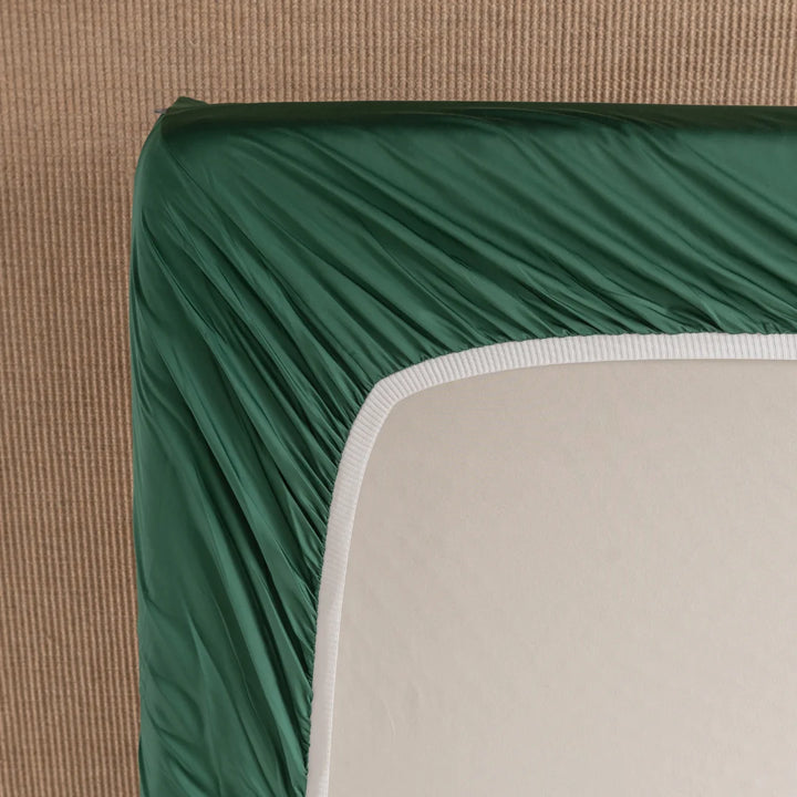 The corner of a mattress is adorned with Linenly's Bamboo Fitted Sheet Set - Evergreen, in a soothing green that offers premium comfort. The elastic edges snugly embrace the off-white mattress, revealing visible stitching as it rests on a textured beige surface.