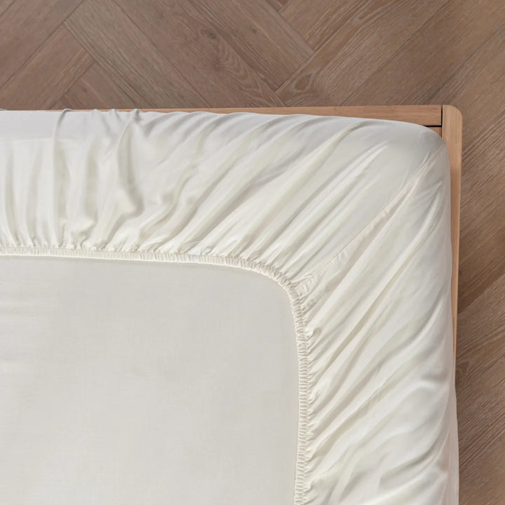A neatly fitted Linenly Ivory Bamboo Fitted Sheet on a corner of a wooden bed frame with a herringbone patterned wooden floor.