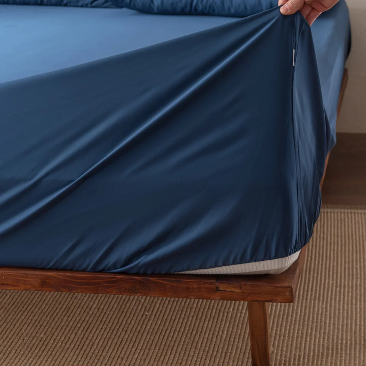 Someone is fitting a Linenly Bamboo Fitted Sheet Set in Midnight over a mattress on a wooden bed frame, adjusting the corner for a smooth finish. A beige rug is partially visible on the floor.