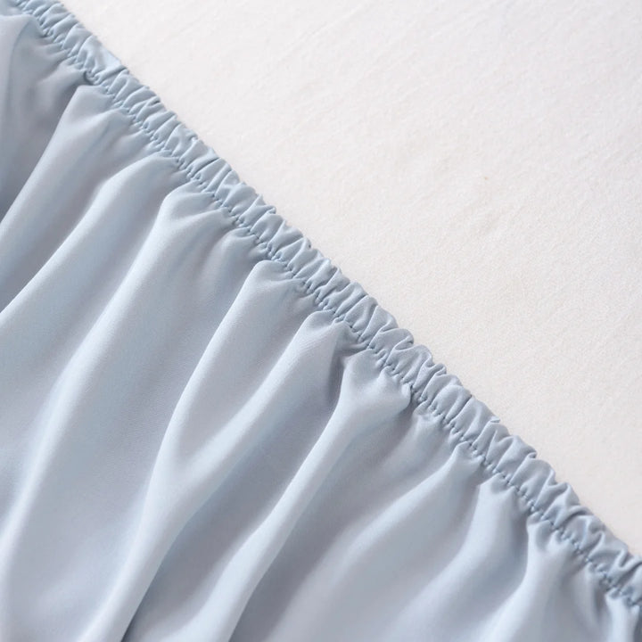 Close-up image of a bed showcasing the Linenly Bamboo Fitted Sheet Set in Pale Blue, with elasticized edges securely fitted over the corner of a mattress. The snug, gathered fit of the eco-friendly sheet provides a cooling feel and stands out against the light-colored mattress.