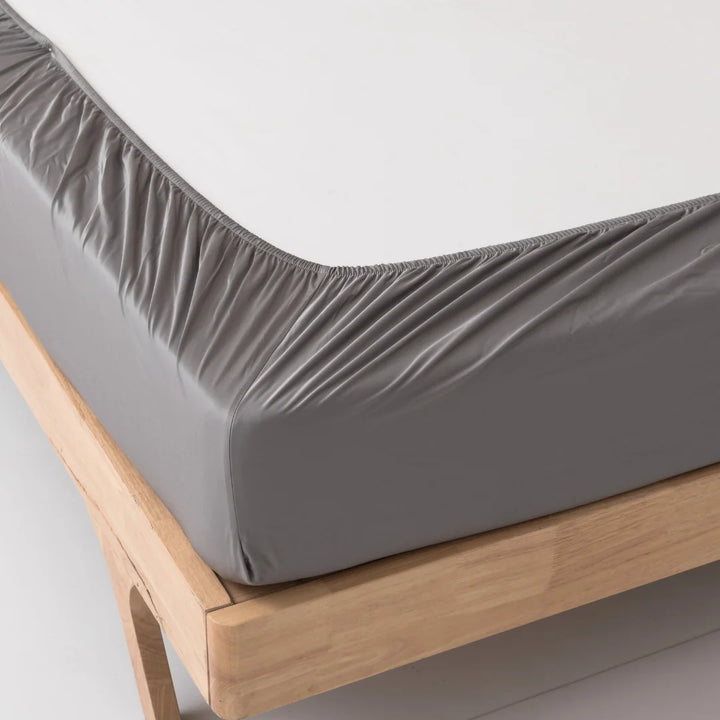 Close-up of a corner of a neatly made bed with a luxuriously soft Linenly Stone Grey Bamboo Fitted Sheet on a wooden bed frame, showcasing sustainability and comfort.
