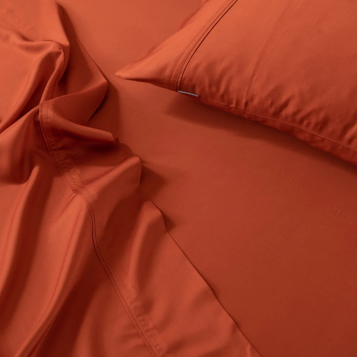 Close-up of a neatly made bed with Linenly's Bamboo Pillowcase Set in Copper Sunset. The rich terracotta fabric appears silky-smooth with subtle stitching, and the organic bamboo sheets have a slightly crinkled texture, adding depth to the image.