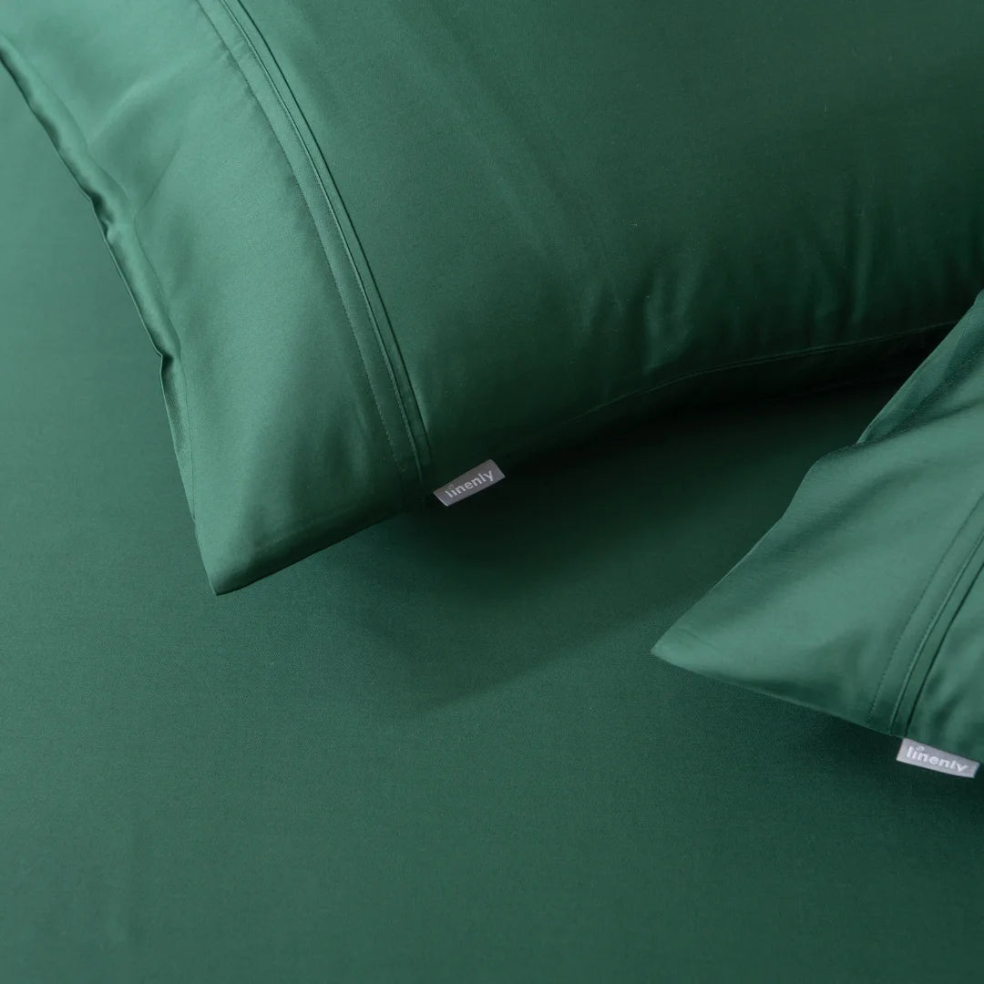 Close-up of a bed featuring Linenly's dark green satin weave sheets and the Bamboo Pillowcase Set in Evergreen. The smooth, well-crafted fabric and visible label enhance a cozy, elegant atmosphere.