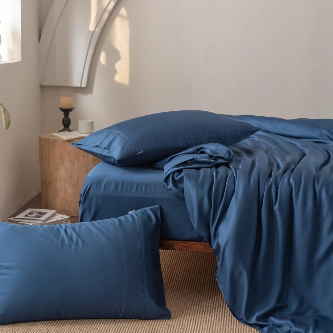 A cozy bedroom features a bed with luxurious blue satin sheets and Linenly's Bamboo Pillowcase Set in Midnight. Sunlight streams in, casting soft shadows, while a wooden bedside table with a candle and plant stands next to a beige rug underneath.