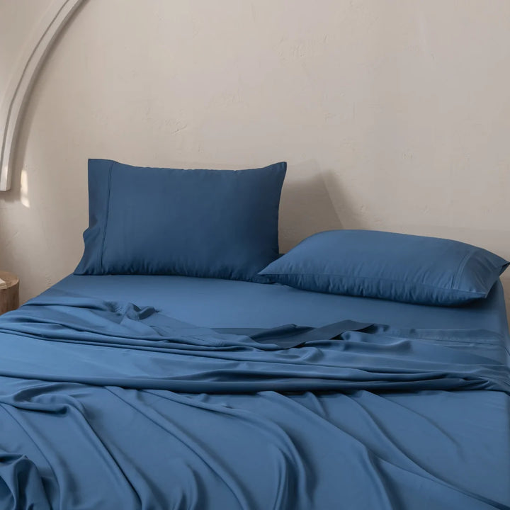 A neatly made bed with blue sheets, Linenly's Bamboo Pillowcase Set - Midnight, and soft natural light streaming over it. The wall is a neutral color that complements the organic bamboo fabric, crafting a calm and inviting atmosphere.