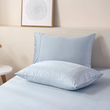 Two fluffy pillows encased in a Linenly organic Bamboo Pillowcase Set in Pale Blue sit atop a neatly made bed with light blue linens against a soft-hued background, creating a serene and inviting atmosphere in a modern bedroom.