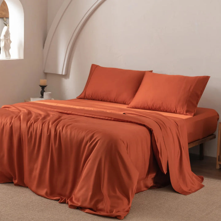 A neatly made bed, draped with a Linenly Bamboo Quilt Cover in Copper Sunset, stands against a white wall featuring a curved arch. A candle and small sculpture decorate the left side table, while beige carpet covers the floor.