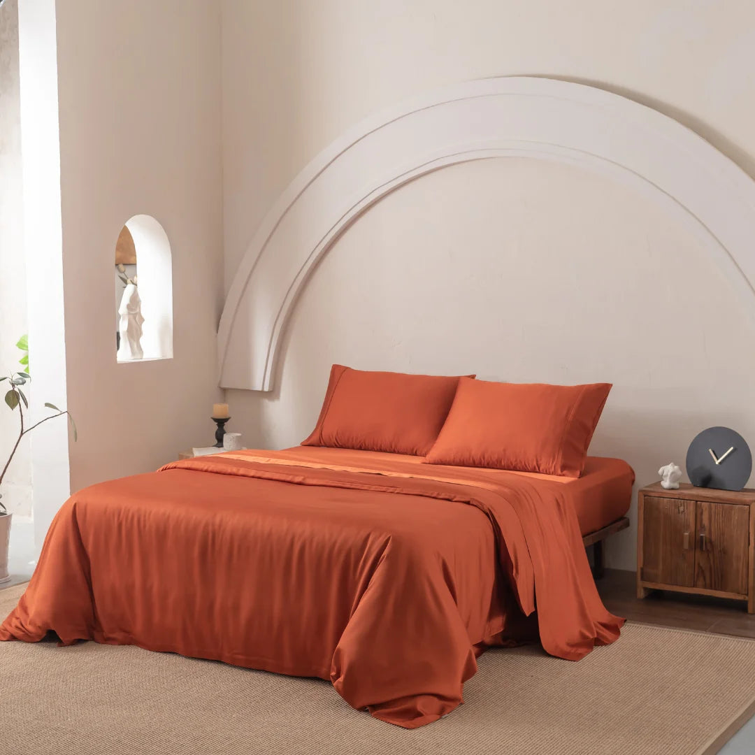 A neatly made bed with Linenly's Bamboo Quilt Cover in Copper Sunset and rust-colored pillows rests against a light beige wall with semi-circular molding. A wooden nightstand holds a modern clock, while soft natural light through an arched window enhances the room's luxurious silkiness.