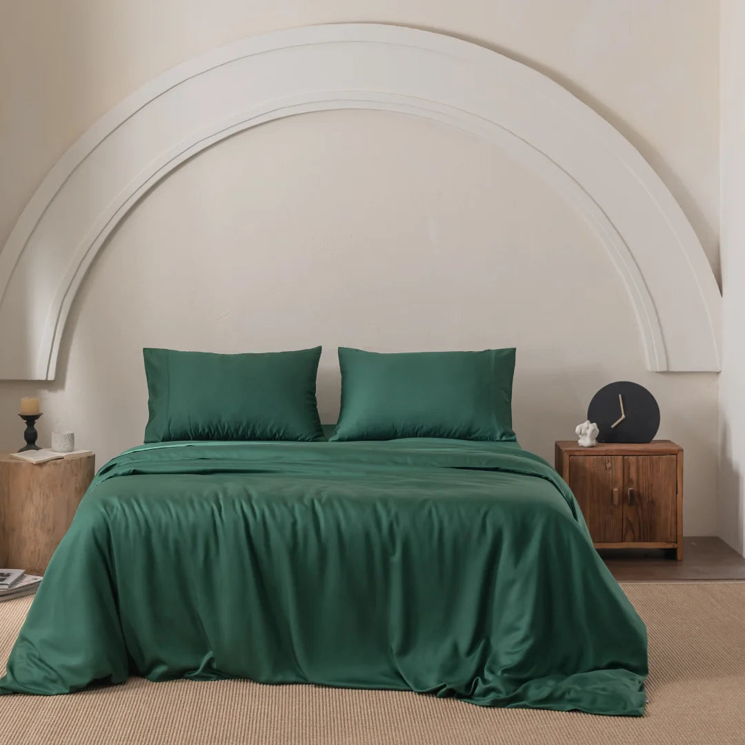 A neatly made bed with Linenly's Bamboo Quilt Cover in Evergreen and two pillows. A wooden nightstand with a clock and candle is on the right, while a wooden stool with a candle is on the left. A white arc adorns the wall above the bed, and a beige rug complements its premium bamboo materials.