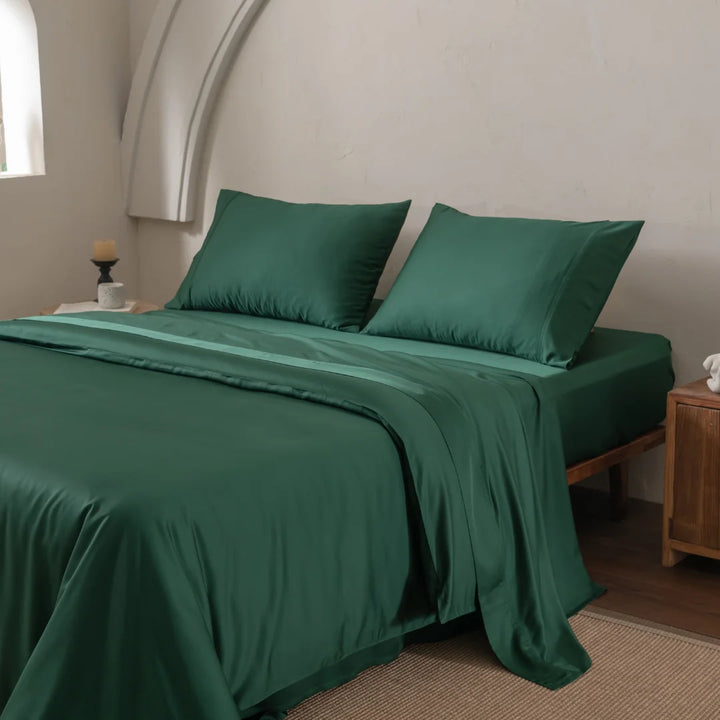 A neatly made bed with Linenly's Bamboo Quilt Cover - Evergreen and dark green sheets, plus two pillows, sits beside a wooden nightstand with a black candle holder. Against an arched white wall, natural light filters in. The sateen weave of the cover exudes luxury.