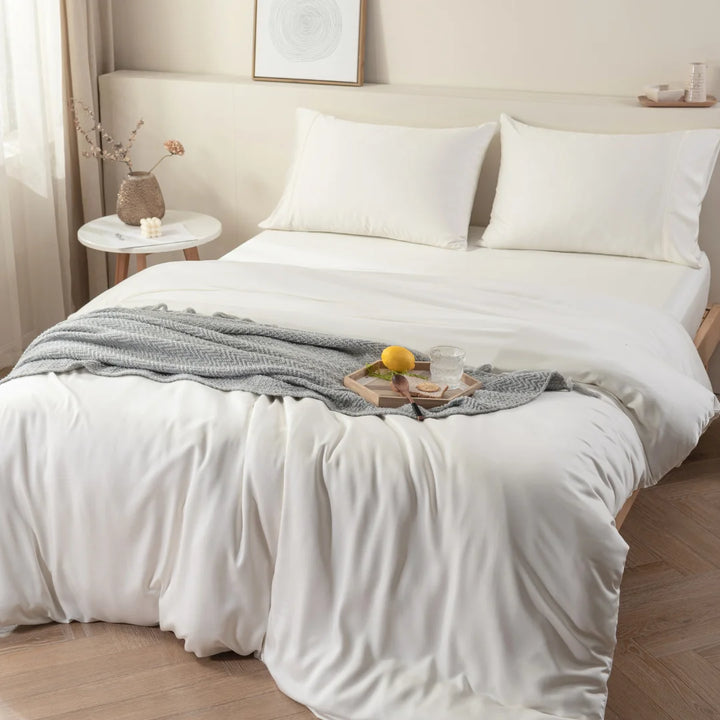 A neatly made bed with a Linenly Ivory Bamboo Quilt Cover in sateen weave, complemented by a textured grey throw at the foot of the bed, and a wooden tray with a book.