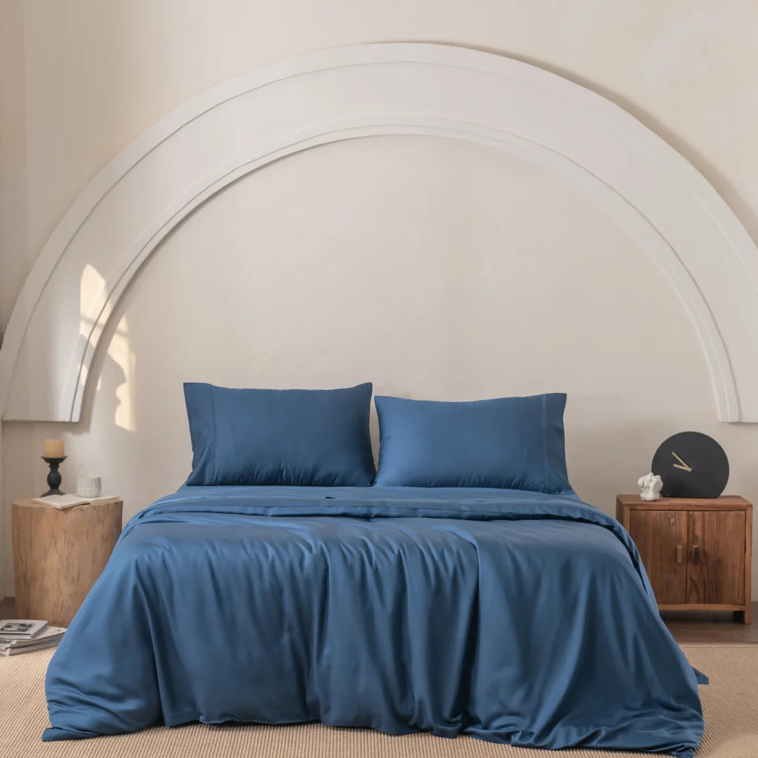 Centered in the room with beige walls and semicircular decor, a bed features two pillows with blue sheets, enveloped in Linenly's Bamboo Quilt Cover Set in Midnight. Wooden nightstands flanking the bed hold decorative items including a candle and clock.