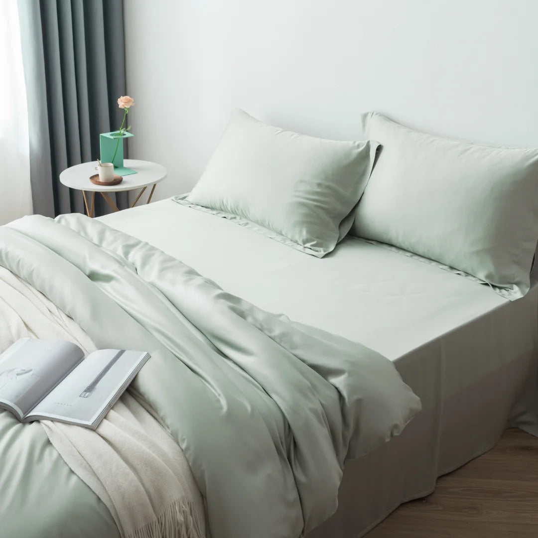 A neatly made bed with a Linenly sage bamboo quilt cover in a minimalist bedroom, accompanied by a small bedside table with a potted plant, and an open book, inviting a peaceful reading session before sleep.