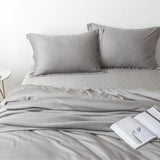 A neatly made bed with grey linens, two pillows, and a Linenly Bamboo Quilt Cover in Stone Grey at the end, accompanied by an open magazine and a small round side table, conveying a minimalist and serene bedroom atmosphere