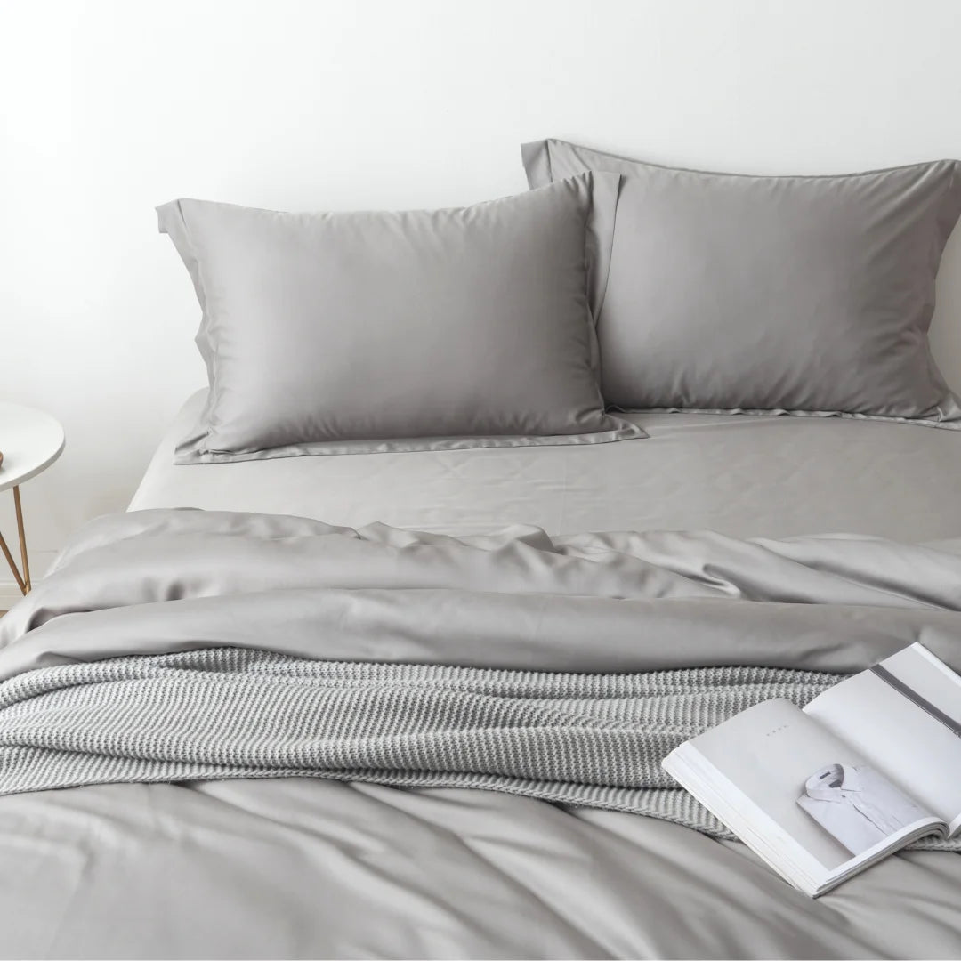 A neatly made bed with grey linens, two pillows, and a Linenly Bamboo Quilt Cover in Stone Grey at the end, accompanied by an open magazine and a small round side table, conveying a minimalist and serene bedroom atmosphere