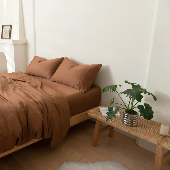 A cozy and minimalist bedroom with a neatly-made bed adorned with a Linenly terracotta bamboo quilt cover made from environmentally friendly bamboo fabric, a wooden bench at the end of the bed holding a potted plant.