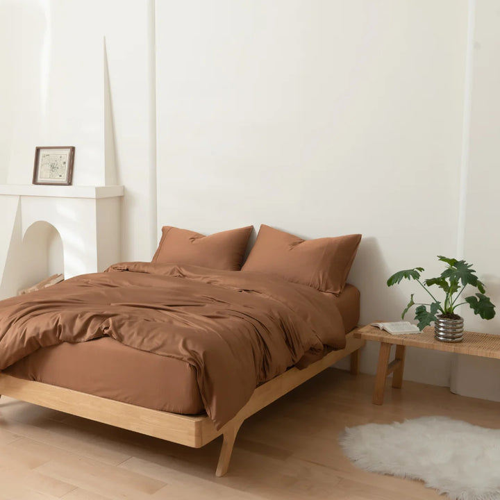 A cozy minimalist bedroom with a low wooden bed, a Linenly Terracotta Bamboo Quilt Cover for both comfort and style, a small bedside table with a plant, and a simple framed picture on the wall.