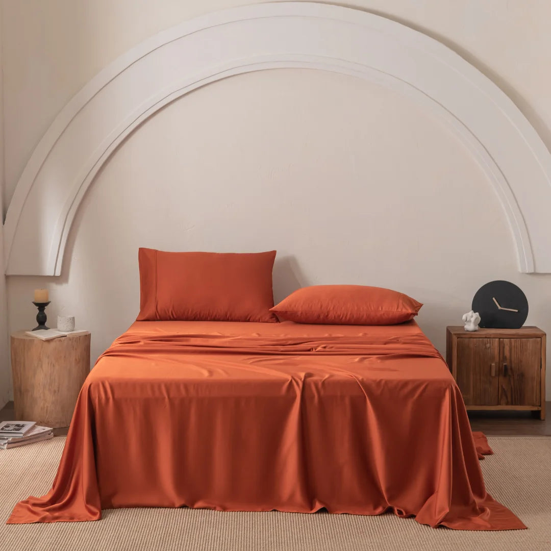 A bed with Linenly's Bamboo Sheet Set in Copper Sunset and two pillows is centered against a beige wall with a large white decorative arch. A wooden nightstand with small decor is on the right, and a wooden stool holding a candle is on the left, all on carpeted flooring.