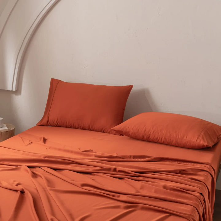 A bed with rumpled Bamboo Sheet Set - Copper Sunset by Linenly and two orange pillows sits against a light beige wall, accompanied by a partially visible round wooden nightstand on the left. The room's arch detail enhances the allure of the ultra-soft, luxurious fabric decor.