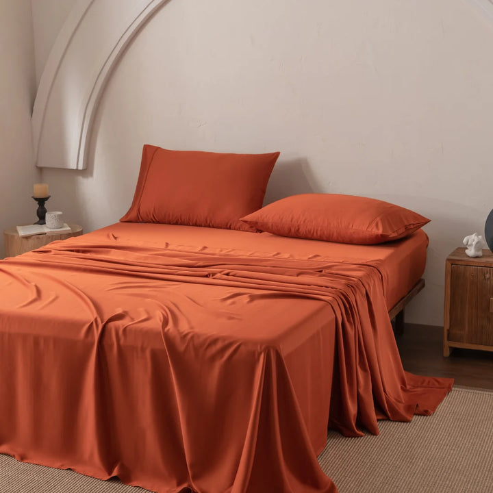 A bedroom features Linenly's Bamboo Sheet Set in Copper Sunset on a bed, luxuriously smooth and slightly draped over the side. The room has a white arched wall, a wooden side table with a candle, and a decorative item on the opposite table.