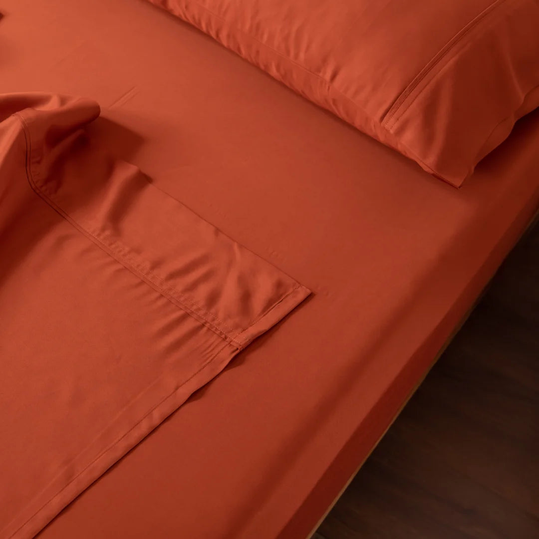 An orange bed with Linenly's Bamboo Sheet Set in Copper Sunset, luxuriously smooth and neatly arranged, features two pillows. The blanket's ultra-soft fabric and detailed stitching are partially revealed, creating an inviting scene on a wooden floor.