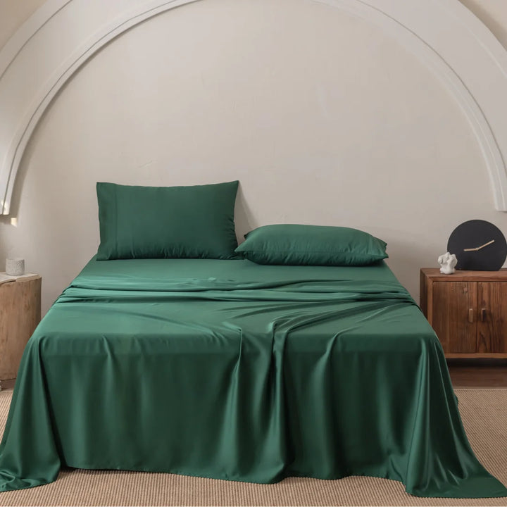 The bed is beautifully arranged with Linenly's Bamboo Sheet Set in Evergreen, featuring ultra-soft, rich green sheets and two pillows. Wooden nightstands are on each side, with a black circular clock on the right, crafting a minimalist and cozy setting against a white arched wall.