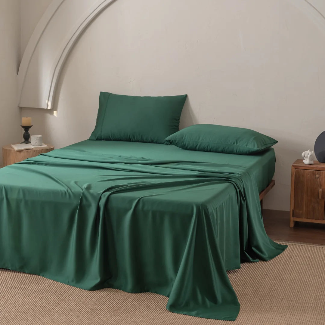 A minimalist room features a neatly made bed with Linenly's Bamboo Sheet Set - Evergreen and two pillows. A candle-topped wooden nightstand and small cabinet flank each side, with a beige rug covering the wooden floor beneath.