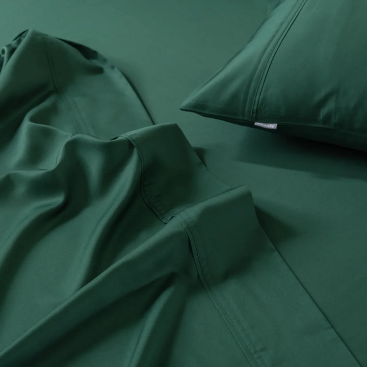 The Bamboo Sheet Set - Evergreen by Linenly features deep green bed linens with a pillowcase and sheets. The silky-smooth texture ensures comfort, and the pillowcase and sheet edges showcase visible stitching. The rich, consistent color enhances the ultra-soft feel.