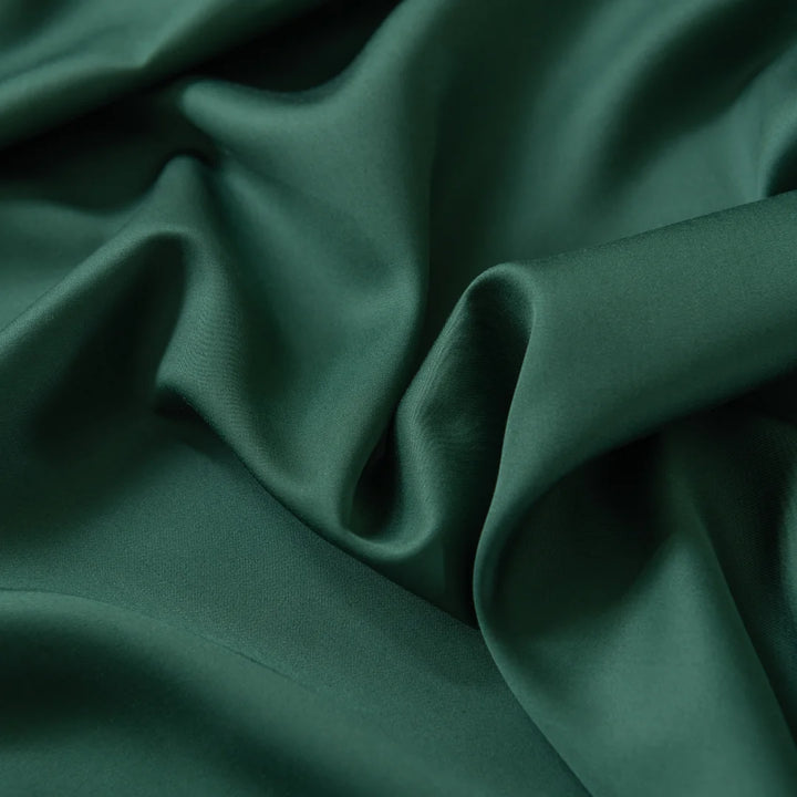 A close-up of dark green satin fabric reveals a silky-smooth texture, gentle folds, and a soft sheen, exuding an ultra-soft and luxurious appearance reminiscent of Linenly's Bamboo Sheet Set - Evergreen.