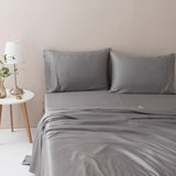 A neatly made bed with a Linenly Stone Grey bamboo sheet set in a minimalist bedroom, complemented by a white lamp and a vase with greenery on a wooden bedside table.