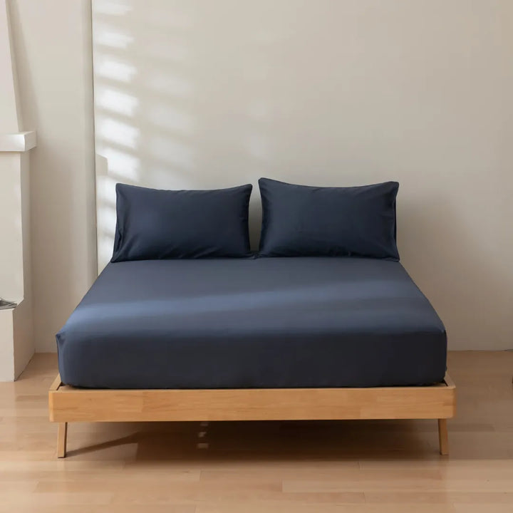 A modern, minimalist bedroom featuring a low-profile wooden bed frame with a Linenly Luxe Sateen Fitted Sheet in Midnight and pillows against a neutral wall for ultimate comfort.