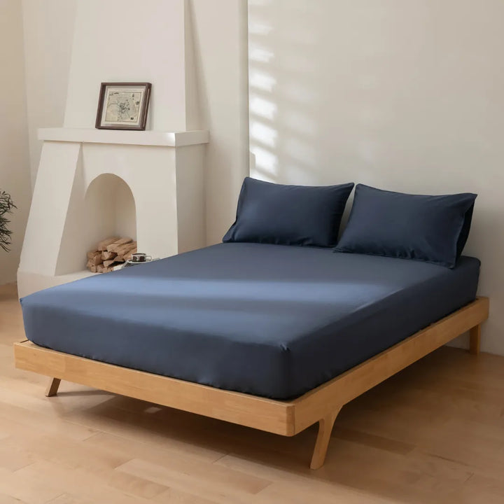 A neatly made bed with a Linenly Luxe Sateen Fitted Sheet in Midnight blue, in a serene room with a white fireplace and subtle decor.