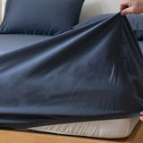 A hand pulls a Linenly Luxe Cotton Sateen Fitted Sheet in Midnight over a mattress on a wooden bed, with matching pillowcases and luxury bedding featured in the background.