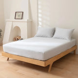 A minimalist bedroom showcases a wooden platform bed with Linenly’s Luxe Cotton Sateen Fitted Sheet in Silver, topped with two pillows, and set against a light wall. Nearby sits a small fireplace with stacked logs and a framed picture.