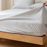 A hand pulls the Linenly Luxe Cotton Sateen Fitted Sheet in Silver over a mattress corner on a light wooden bed, with white pillows and bedding in a sunlit room.