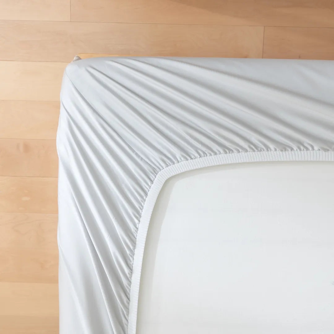 Close-up of the Luxe Cotton Sateen Fitted Sheet - Silver by Linenly, partially pulled over a mattress, showcasing its elastic edge and premium details against a wood floor backdrop.