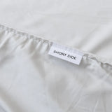 The Linenly Luxe Cotton Sateen Fitted Sheet in Silver is crafted from long-staple cotton, with a sewn seam and a small white "SHORT SIDE" label in black text to mark the short edge—perfect for luxury bedding and easy placement.