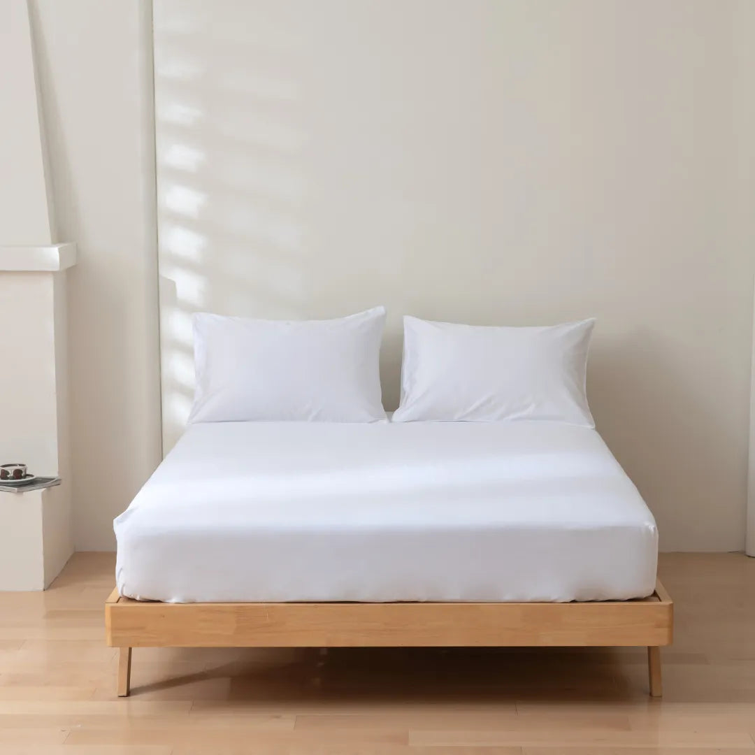 A minimalist bedroom with a wooden platform bed, featuring Linenly’s Luxe Cotton Sateen Fitted Sheet in white and two white pillows, set against a plain light-colored wall and a light wood floor.