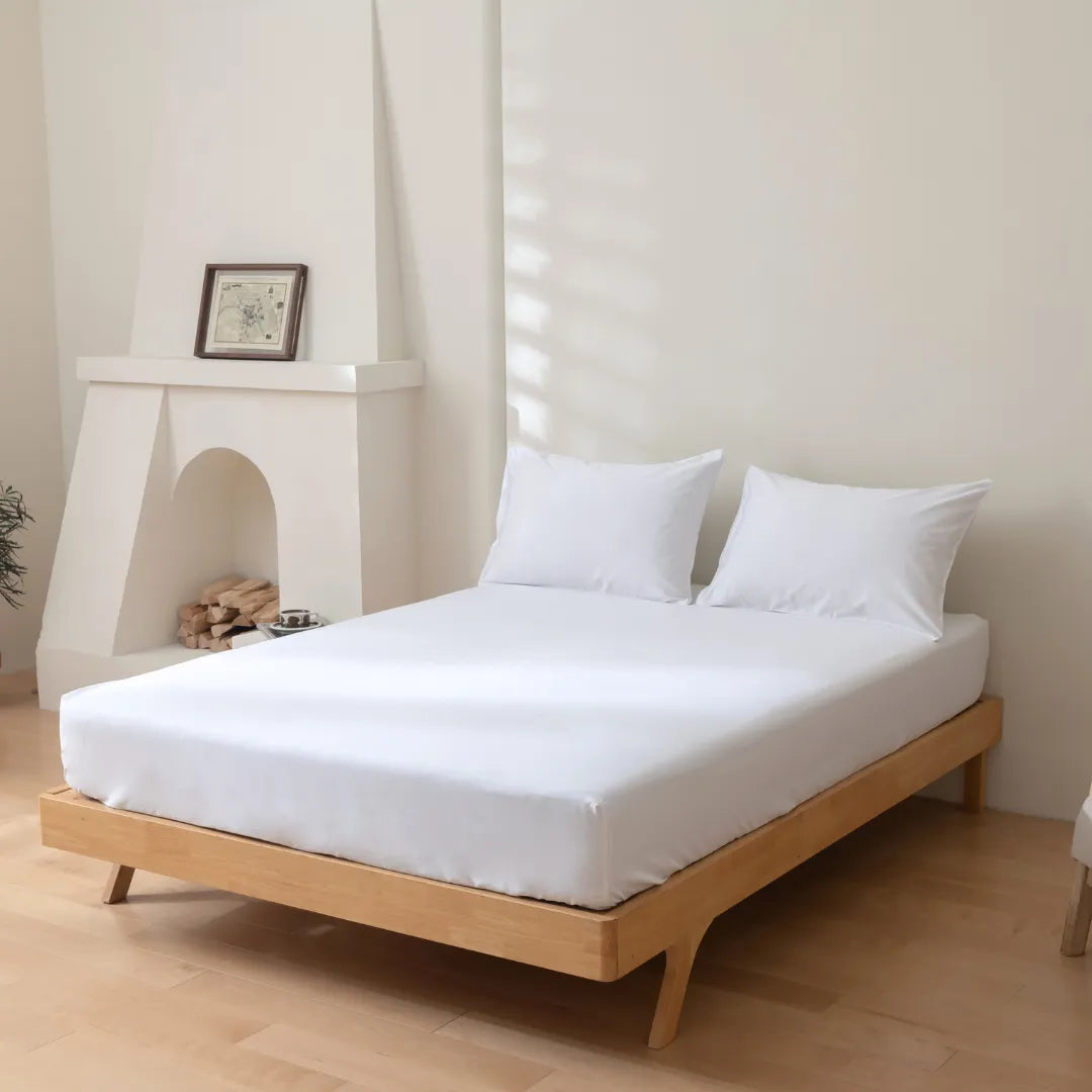 A minimalist bedroom featuring a low-profile wooden bed dressed in Linenly Luxe Sateen Fitted Sheets in white, flanked by a decorative fireplace with a framed picture atop, set against a neutral wall, creating a serene and uncluttered vibe.