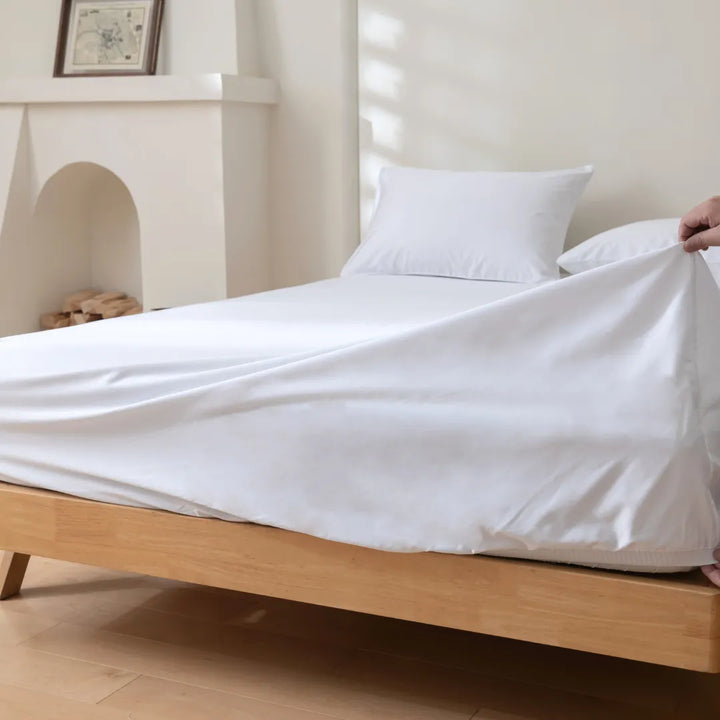 A freshly made bed in a bright room with a person tucking in Linenly Luxe Sateen Fitted Sheet in white cotton.