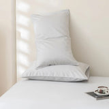 Two white pillows rest on a neatly made bed with the Linenly Luxe Cotton Sateen Flat Sheet in Silver. A cup and saucer sit on a tray nearby as soft natural light casts gentle shadows across the room.