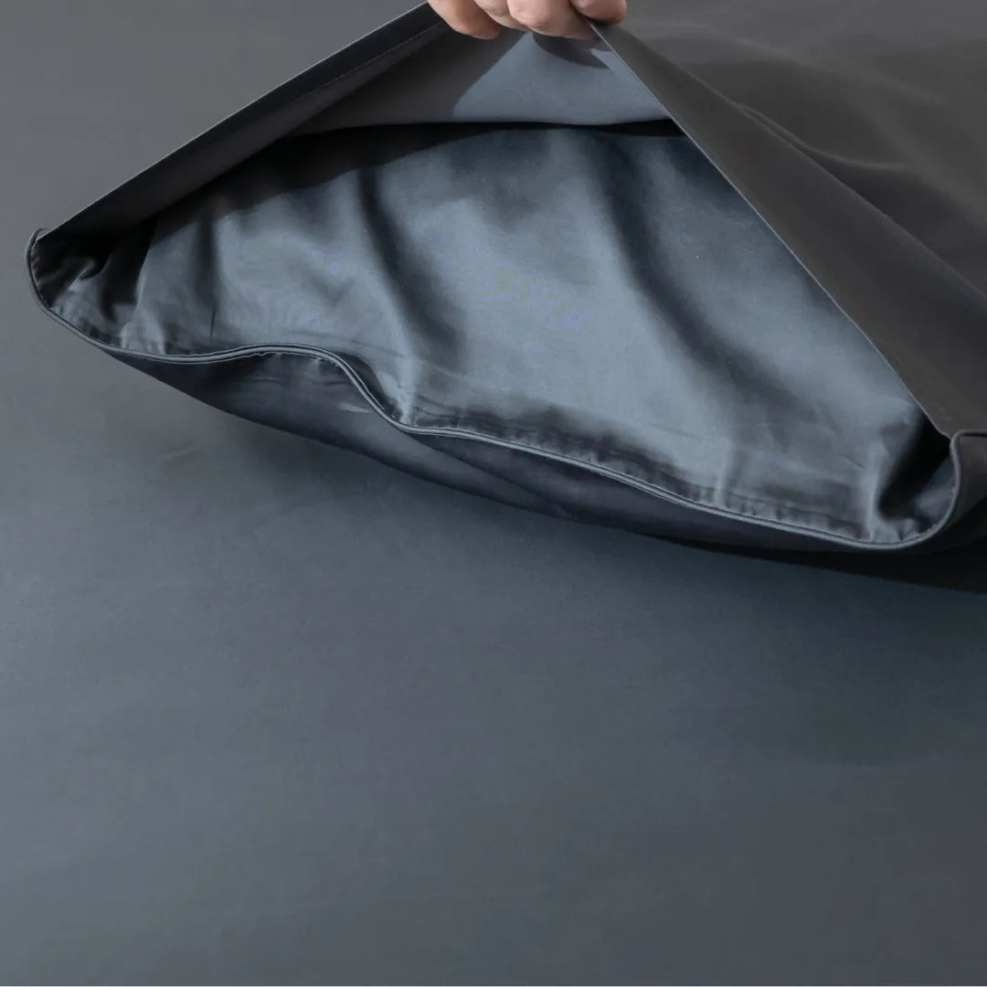 A hand lifts the flap of a Linenly Luxe Cotton Sateen Pillowcase in charcoal, revealing a pillow inside, on a matching dark gray surface.