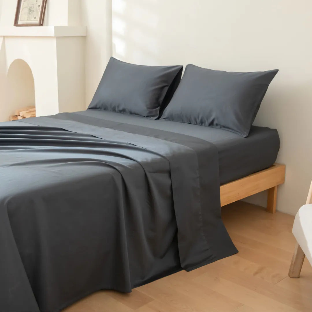 A bed is styled with Linenly's Luxe Cotton Sateen Pillowcase Set in Charcoal, paired with dark gray sheets, all set on a light wooden frame in a bright, minimalist room with pale walls and wood floors.