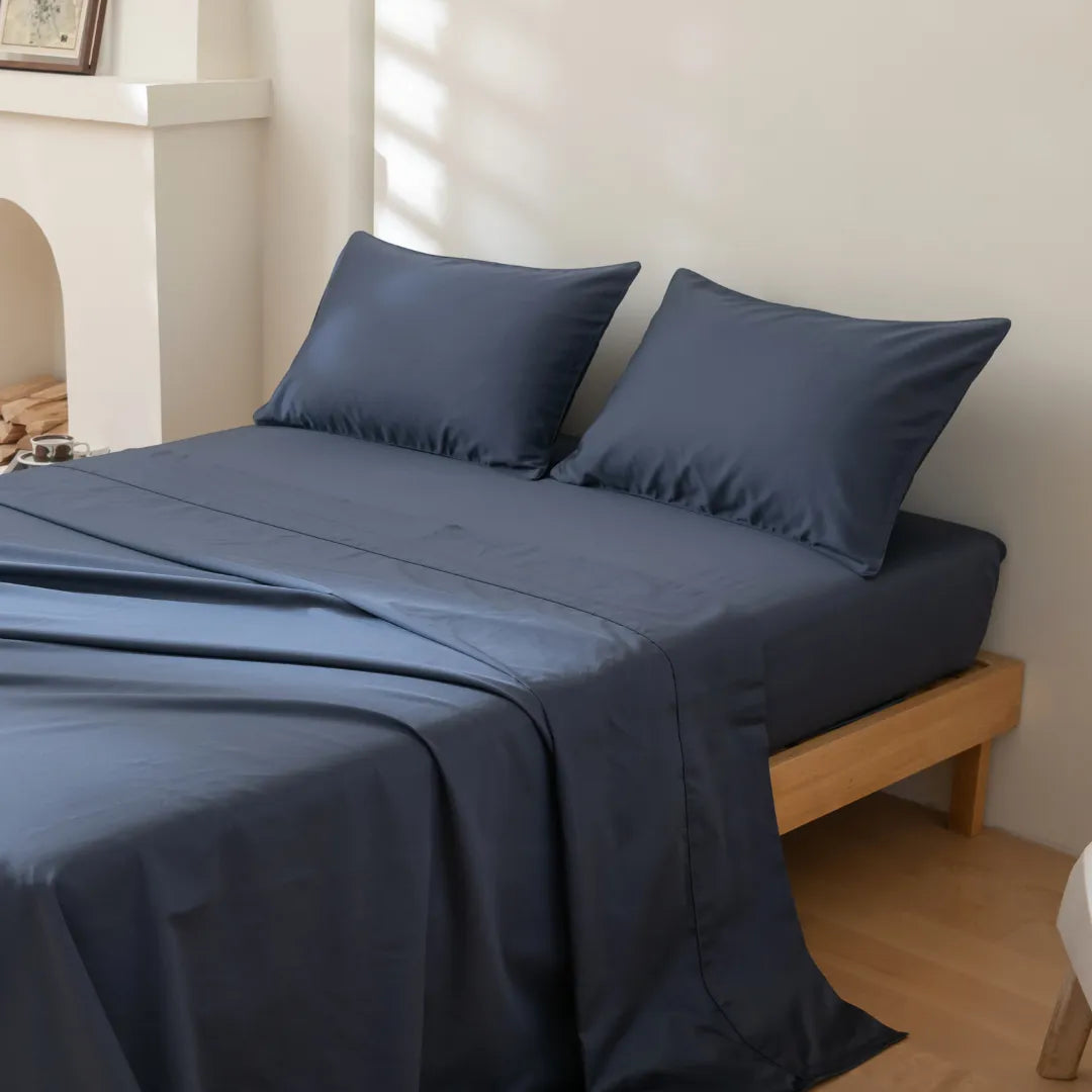 A neatly made bed features the Linenly Luxe Cotton Sateen Pillowcase Set in Midnight, with two matching pillows on a simple wooden frame in a bright, minimalist bedroom as sunlight streams in and casts soft shadows on the bed.